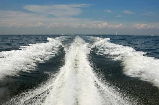 A wake seen from a stern, the water thrown white, the coastline far behind β the frame mostly spray.