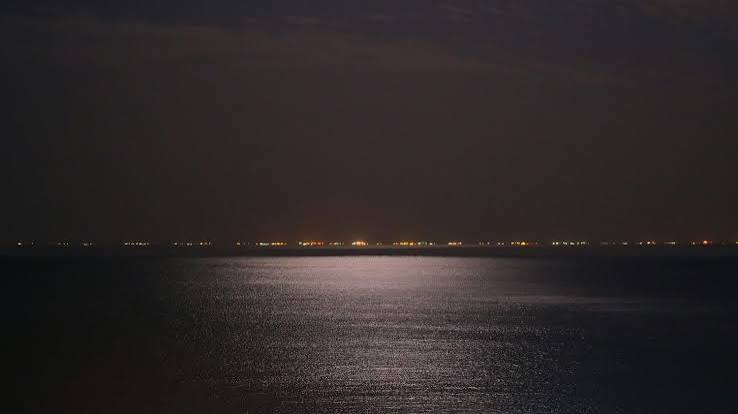 A coastline at night, distant fires along the shore over dark water, slightly out of focus.