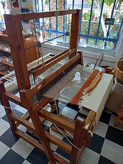 A wooden loom mid-weave by a window, nobody at it.